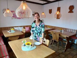 Nicole Ehmer steht lächelnd im hellen Frühstücksraum mit Holztischen, Eckbänken und Holz-Pendelleuchten