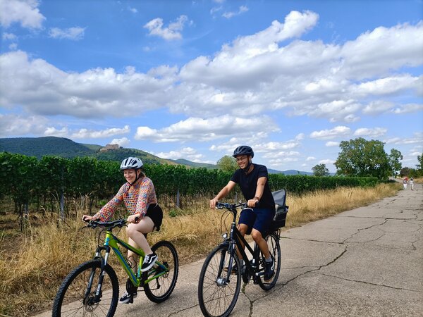 Theresa und Daniel Hoefer fahren mit Fahrradhelm auf einem Wirtschaftsweg durch die Weinberge, im Hintergrund das Hambacher Schloss
