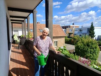 Marianne Ehmer gießt mit einer grünen Gießkanne die Blumen auf dem langen Holzbalkon des Gästehauses