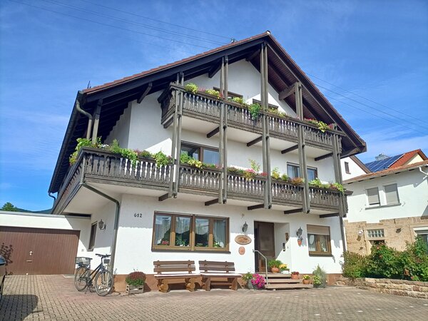 Weißes Gästehaus mit zwei umlaufenden Holzbalkonen, Blumenkästen, Sitzbänken und Fahrrad vor dem Eingang