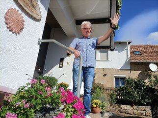 Matthias Ehmer winkt lachend von der Eingangstreppe des Gästehauses, davor blühende Geranien