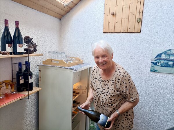Marianne Ehmer nimmt lächelnd eine Weinflasche aus dem Getränkekühlschrank, daneben Weinflaschen und Gläser im Regal